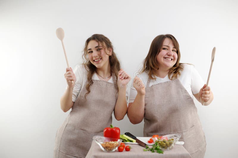 2 women dancing while preparing tasty healthy food vegetarian food vegetable salad in hands wooden spoons identical kitchen aprons white background joy fun family. 2 women dancing while preparing tasty healthy food vegetarian food vegetable salad in hands wooden spoons identical kitchen aprons white background joy fun family