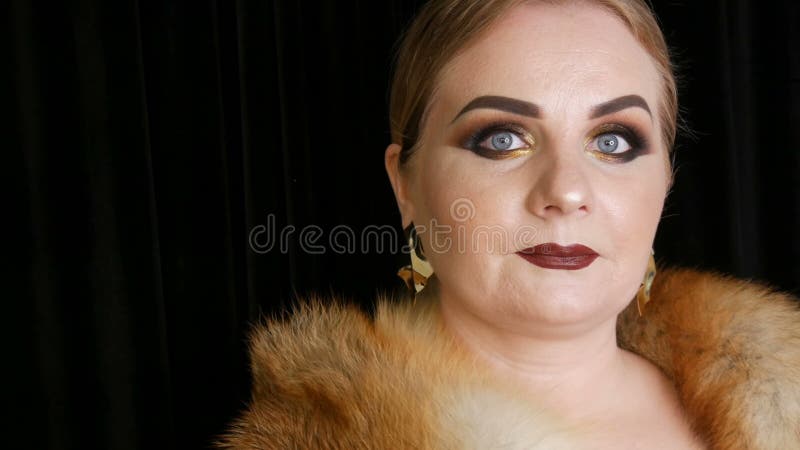 Жирная женская модель с золотым макияжем и закоптелые глаза в пальто меха лисы и больших золотых серег представляя на черноте видеоматериал