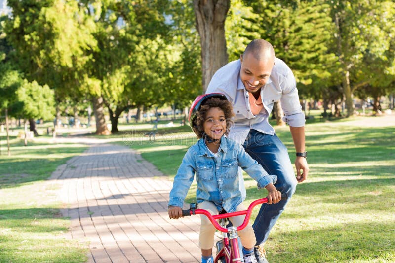 Lovely father teaching son riding bike at park. Happy father helping excited son to ride a bicycle in a summer day. Young smiling black boy wearing bike helmet while learning to ride cycle with his dad. Lovely father teaching son riding bike at park. Happy father helping excited son to ride a bicycle in a summer day. Young smiling black boy wearing bike helmet while learning to ride cycle with his dad.