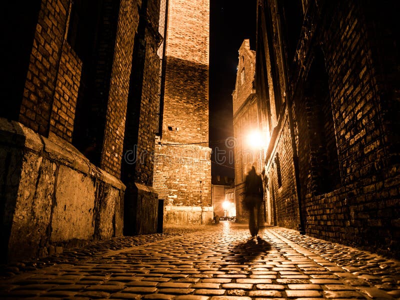 Illuminated cobbled street with light reflections on cobblestones in old historical city by night. Dark blurred silhouette of person evokes Jack the Ripper. Illuminated cobbled street with light reflections on cobblestones in old historical city by night. Dark blurred silhouette of person evokes Jack the Ripper.