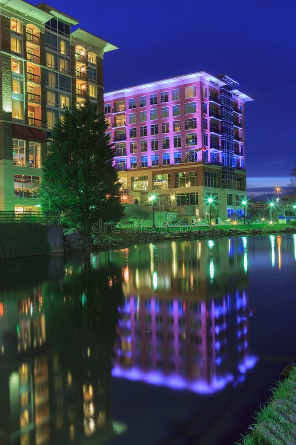 Buildings illuminated at night with green and purple lights and reflected in the Reedy River in downtown Greenville, South Carolina. Buildings illuminated at night with green and purple lights and reflected in the Reedy River in downtown Greenville, South Carolina.