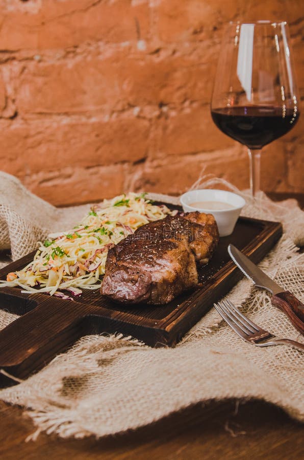 Зажаренный стейк говядины ribeye с капустой тушёного мяса и рюмкой красного вина, трав и специй над коричневой дерюгой на деревян стоковые изображения rf Зажаренный стейк говядины ribeye с капустой тушёного мяса и рюмкой красного вина, трав и специй над коричневой дерюгой на деревян стоковые изображения rf