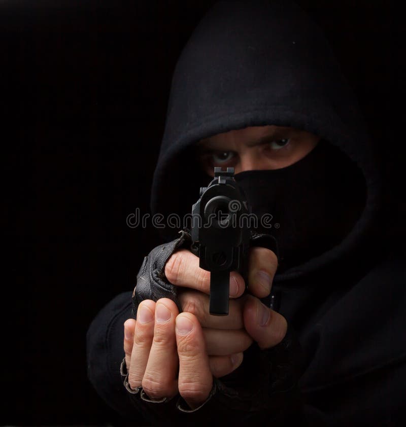 Masked robber with gun aiming into the camera against a black background. Masked robber with gun aiming into the camera against a black background