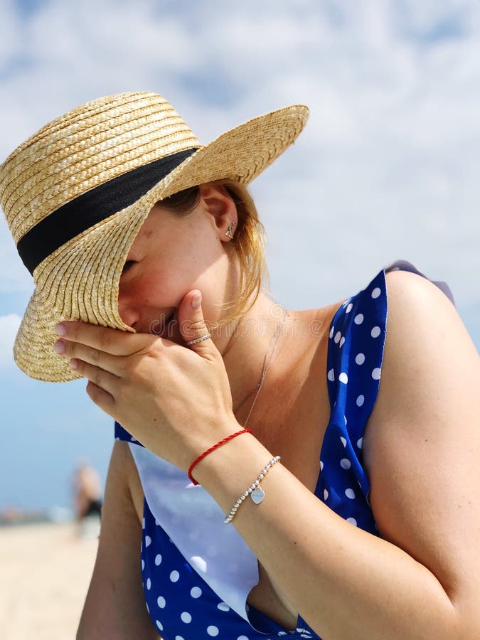 Young beautiful woman in summer hat, laughs with shy, in blue dress with dots at summer beach. Young beautiful woman in summer hat, laughs with shy, in blue dress with dots at summer beach