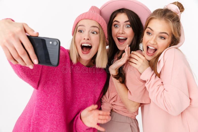 Image of three beautiful girls wearing pink clothes smiling and taking selfie photo on cellphones isolated over white background. Image of three beautiful girls wearing pink clothes smiling and taking selfie photo on cellphones isolated over white background