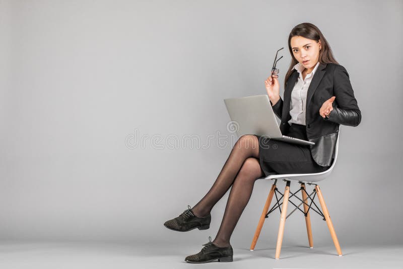 Изображение молодой бизнес-женщины, позирующей изолированную на фоне серой стены сидящей на стуле с помощью компьютера