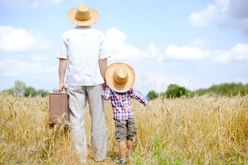 Father and his son in straw hats travelling on summer wheat field. Man carrying a heavy suitcase and holding child's hand. They both are looking in the distance. The sun is shining. Father and his son in straw hats travelling on summer wheat field. Man carrying a heavy suitcase and holding child's hand. They both are looking in the distance. The sun is shining.