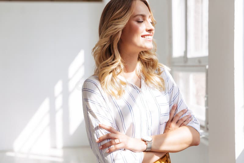Image of joyful elegant blonde woman smiling and standing with arms crossed in white office. Image of joyful elegant blonde woman smiling and standing with arms crossed in white office