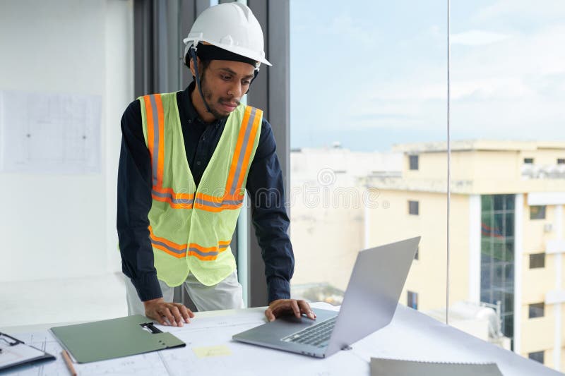 Serious construction engineer in vest and hardhat checking e-mails on laptop screen. Serious construction engineer in vest and hardhat checking e-mails on laptop screen