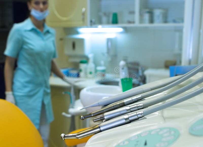 The focus is placed on the tools of the dentist (drills), while the room and the nurse in the background are out of focus. The focus is placed on the tools of the dentist (drills), while the room and the nurse in the background are out of focus.