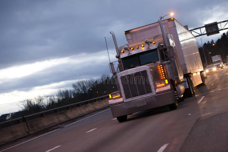 A powerful big rig semi truck with a full-sized dry van trailer for the transport of industrial goods, with high exhaust pipes and a lot of marker lights moving along the evening highway against the background of a cloudy sky. A powerful big rig semi truck with a full-sized dry van trailer for the transport of industrial goods, with high exhaust pipes and a lot of marker lights moving along the evening highway against the background of a cloudy sky