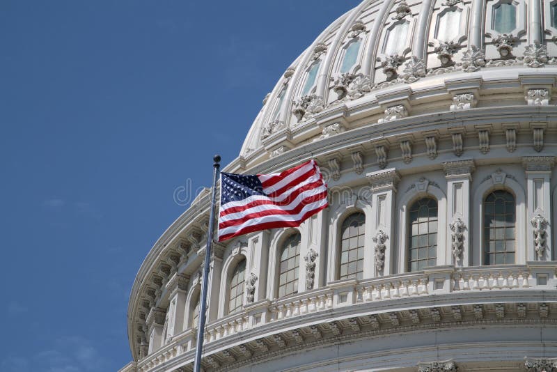 The dome of the US United States Capitol building for Democrat Republican Government Senate and House congress parties under a summer blue sky with the American flag in front of it. The dome of the US United States Capitol building for Democrat Republican Government Senate and House congress parties under a summer blue sky with the American flag in front of it.