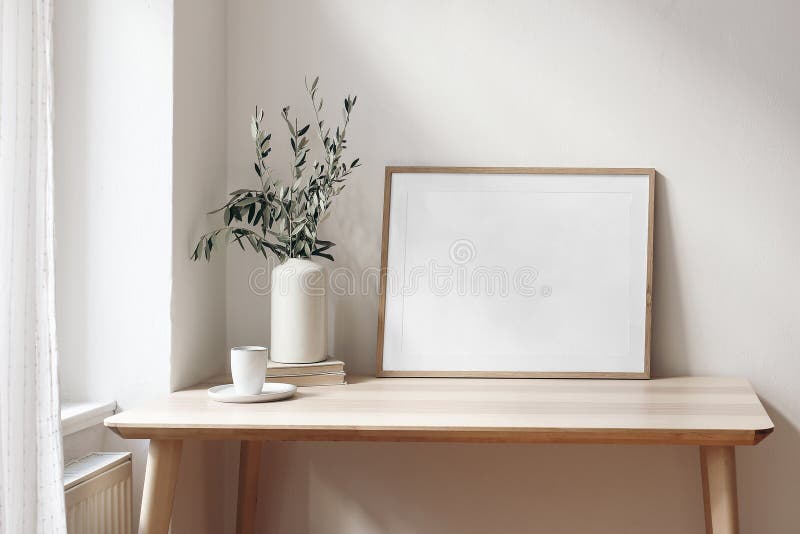 Home office concept. Empty horizontal wooden picture frame mockup. Cup of coffee on wooden table. White wall background. Vase with olive branches, elegant working space. Scandinavian interior design. Home office concept. Empty horizontal wooden picture frame mockup. Cup of coffee on wooden table. White wall background. Vase with olive branches, elegant working space. Scandinavian interior design.