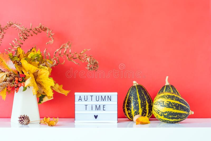 Коробка с текстом AUTUMN TIME, декоративные полосатые тыквы, ваза с букетом падающих листьев и паром на фоне ярко-красной стены стоковое фото