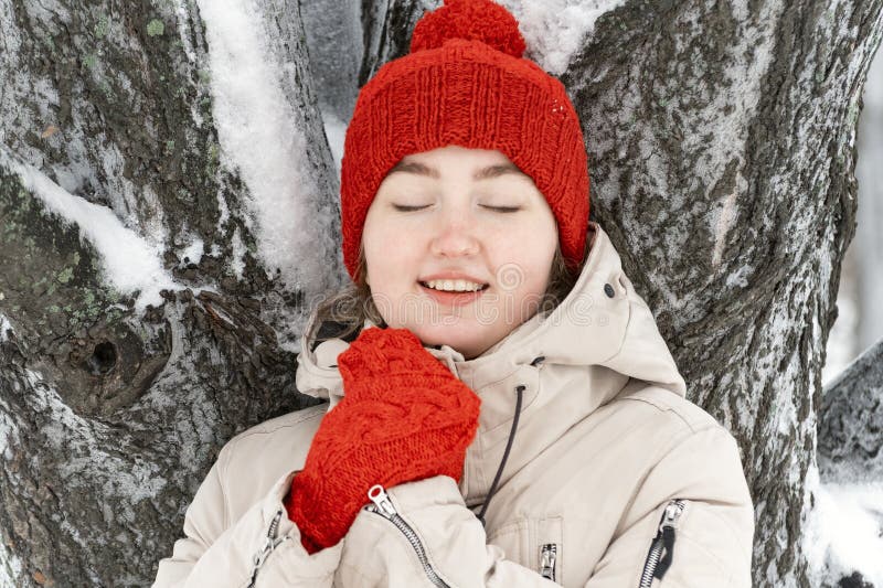 красивая девушка улыбается с закрытыми глазами и стоит на заснеженном дереве. женщина в модной красной шляпе и варежки зимой стоковые изображения