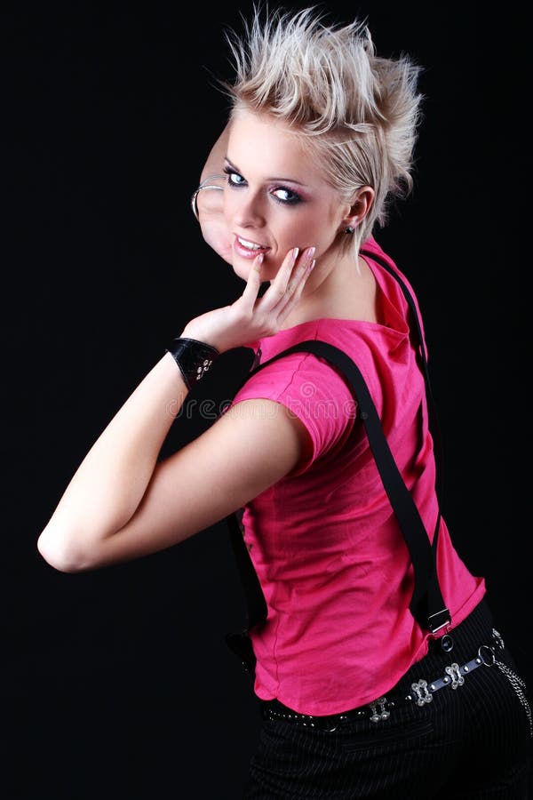 Beautiful young girl with a modern spiky blond hairstyle wearing a vivid pink top and braces having fun at a party standing looking back at the camera with her hands raised on a black background. Beautiful young girl with a modern spiky blond hairstyle wearing a vivid pink top and braces having fun at a party standing looking back at the camera with her hands raised on a black background