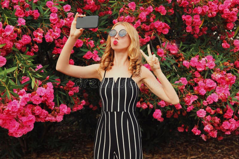 Beautiful young woman taking selfie picture by smartphone blowing red lips sending sweet air kiss over flowers roses background. Beautiful young woman taking selfie picture by smartphone blowing red lips sending sweet air kiss over flowers roses background