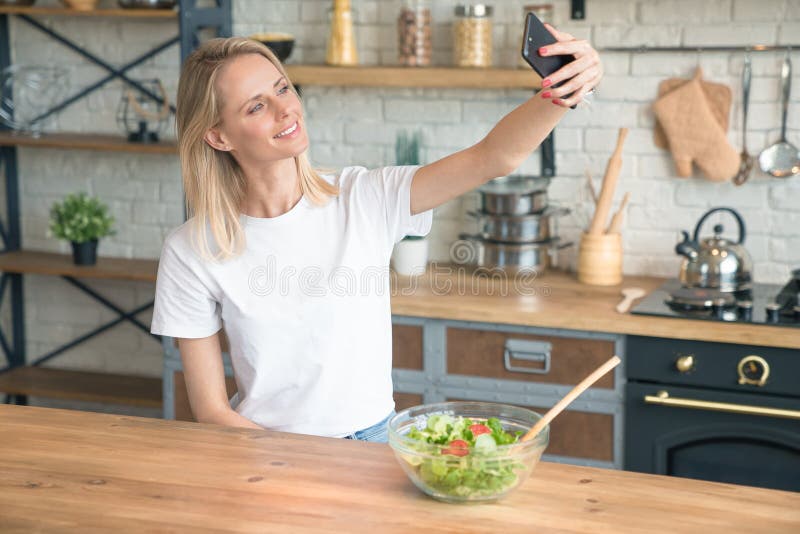 Красивая молодая усмехаясь женщина делая selfie с телефоном пока делающ салат в кухне E салат овоща E стоковое фото Красивая молодая усмехаясь женщина делая selfie с телефоном пока делающ салат в кухне E салат овоща E стоковое фото
