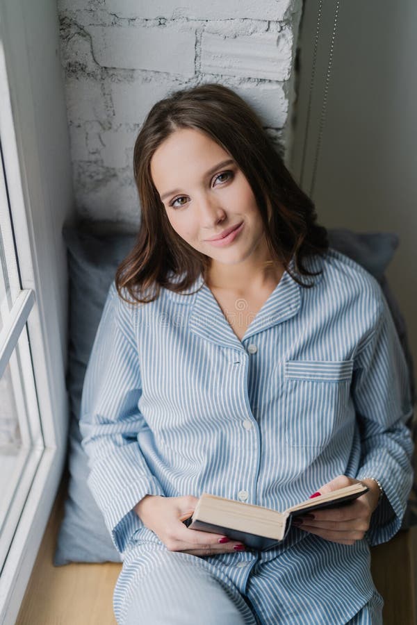 Красивая расслабленная женщина при беспечальное выражение, одетое в nightclothes, читает книгу на силле окна в утре, наслаждается стоковое изображение rf Красивая расслабленная женщина при беспечальное выражение, одетое в nightclothes, читает книгу на силле окна в утре, наслаждается стоковое изображение rf