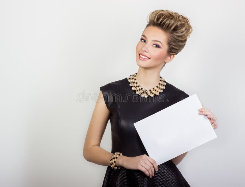 Portrait of a beautiful happy young woman smiling in a black evening dress with hair and make-up with expensive jewelry with a white sign in his hands in Studio on white background. Portrait of a beautiful happy young woman smiling in a black evening dress with hair and make-up with expensive jewelry with a white sign in his hands in Studio on white background