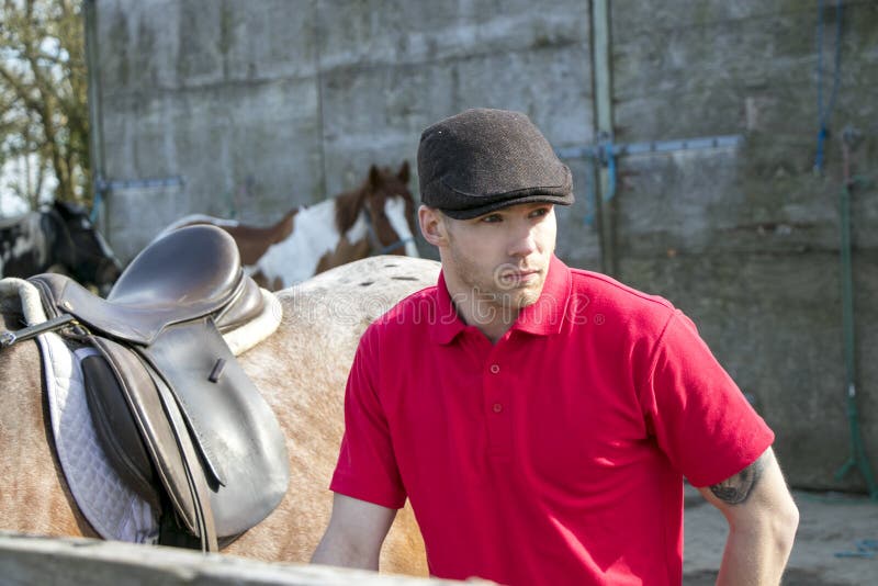Красивый всадник лошади в красной рубашке поло и flatcap стоя рядом с его оседланной лошадью стоковые фотографии rf Красивый всадник лошади в красной рубашке поло и flatcap стоя рядом с его оседланной лошадью стоковые фотографии rf