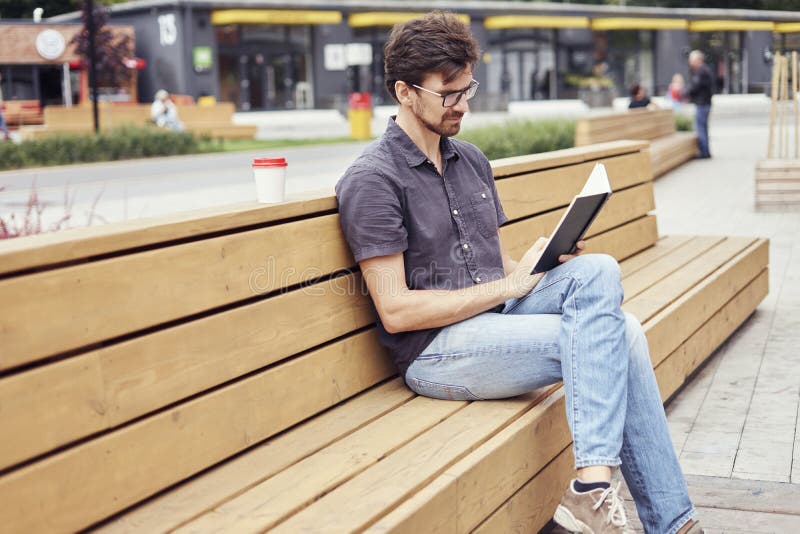 Handsome man reading book sitting outside in public space. Wearing glasses alone working. Concept of education students. Handsome man reading book sitting outside in public space. Wearing glasses alone working. Concept of education students.