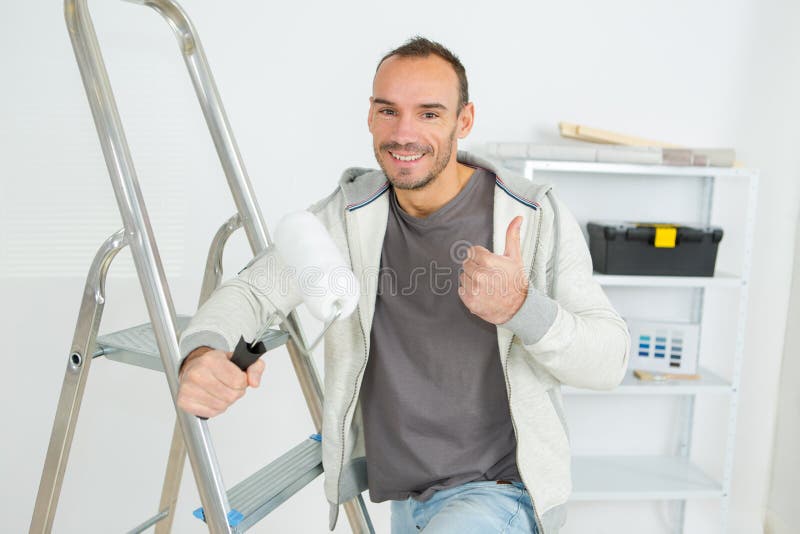 Красивый молодой decorator с валиком краски, делая большие пальцы руки жест стоковая фотография