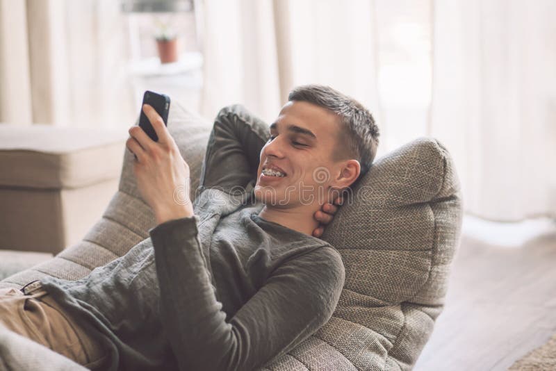 Handsome teenage guy with dental braces smiling and using smartphone while relaxing on modern couch in living room interior. Happy man resting in soft chair at home. Handsome teenage guy with dental braces smiling and using smartphone while relaxing on modern couch in living room interior. Happy man resting in soft chair at home