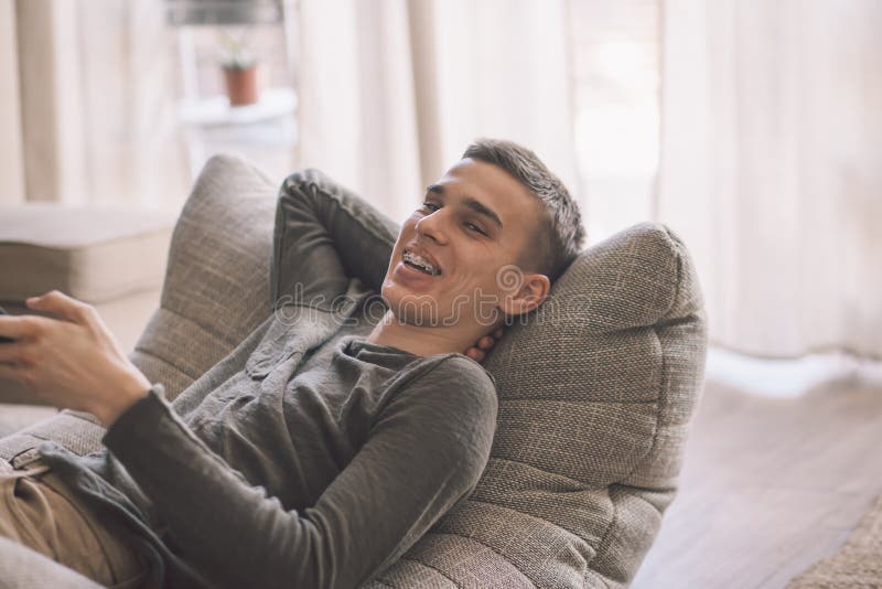 Handsome teenage guy with dental braces smiling while relaxing on modern couch in living room interior. Happy man resting in soft chair at home. Handsome teenage guy with dental braces smiling while relaxing on modern couch in living room interior. Happy man resting in soft chair at home