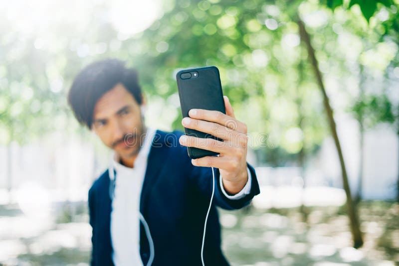 Красивый усмехаясь бизнесмен используя smartphone для listining музыки пока идущ в парк города Молодой человек делая selfie стоковое фото Красивый усмехаясь бизнесмен используя smartphone для listining музыки пока идущ в парк города Молодой человек делая selfie стоковое фото