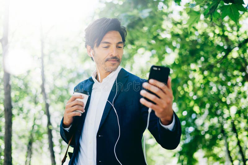 Красивый усмехаясь бизнесмен используя smartphone для listining музыки пока идущ в парк города Молодой человек делая selfie стоковая фотография rf Красивый усмехаясь бизнесмен используя smartphone для listining музыки пока идущ в парк города Молодой человек делая selfie стоковая фотография rf