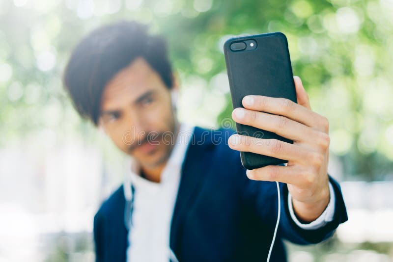 Красивый усмехаясь бизнесмен используя smartphone для listining музыки пока идущ в парк города Молодой человек делая selfie стоковая фотография Красивый усмехаясь бизнесмен используя smartphone для listining музыки пока идущ в парк города Молодой человек делая selfie стоковая фотография