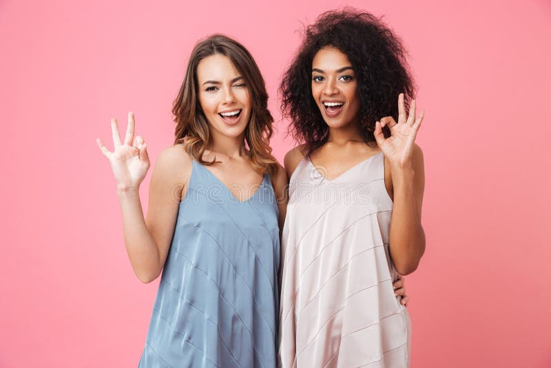 Two beautiful summer girls 20s with different color of skin in dresses smiling at camera and showing ok or alright sign isolated over pink background. Two beautiful summer girls 20s with different color of skin in dresses smiling at camera and showing ok or alright sign isolated over pink background