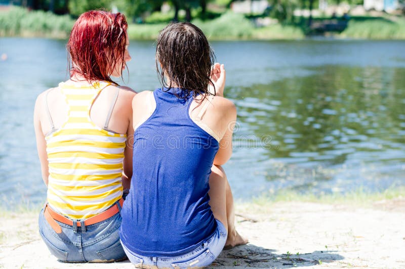 Two young pretty women in wet clothing shirts having fun relaxing sitting on the bank of the river on sandy beach ground and talking together on summer green outdoors. Two young pretty women in wet clothing shirts having fun relaxing sitting on the bank of the river on sandy beach ground and talking together on summer green outdoors
