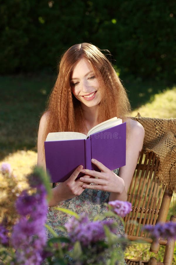 Красная с волосами женщина в саде лета читая книгу стоковое изображение