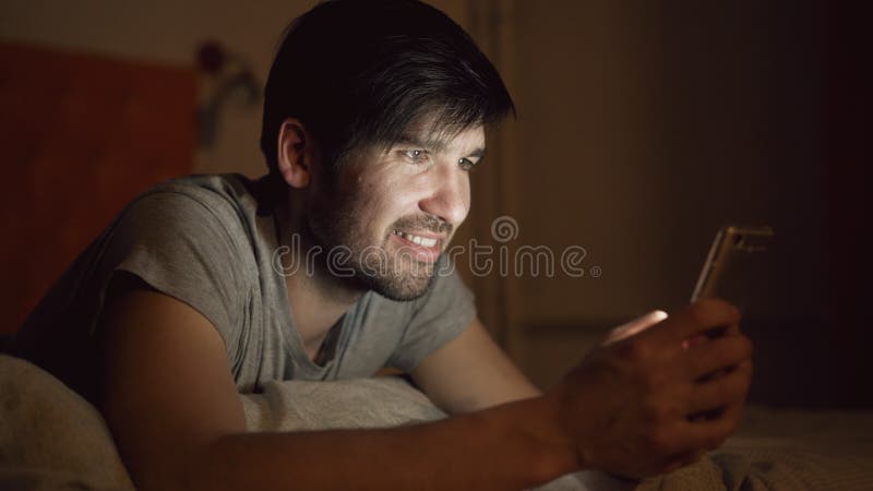 Крупный план молодого усмехаясь человека используя smartphone для серфинга социальных средств массовой информации лежа в кровати стоковое изображение rf