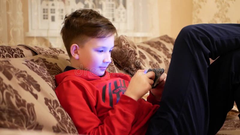 11-летний мальчик сидит на диване и играет на смартфоне. видеоматериал - Видео насчитывающей технология, счастливо: 215332610