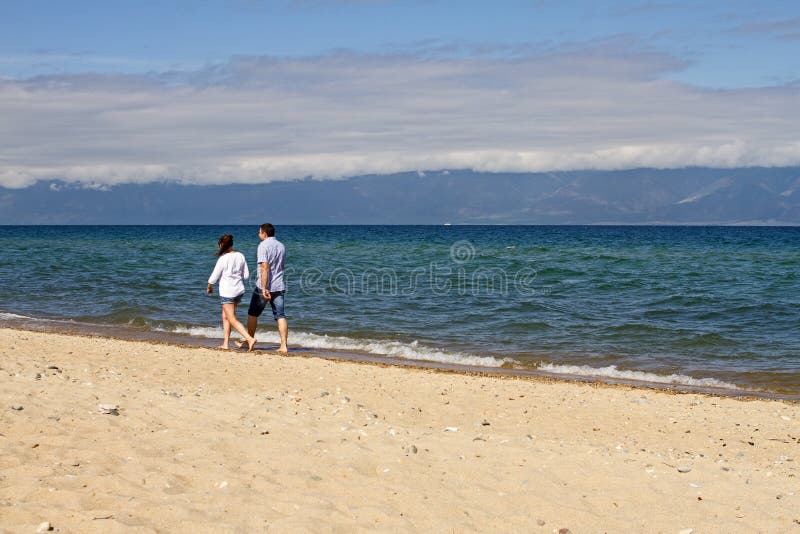 Любящая пара человека и женщины идет вдоль береговой линии песка Lake Baikal на предпосылке горы на солнечный день стоковое изображение rf Любящая пара человека и женщины идет вдоль береговой линии песка Lake Baikal на предпосылке горы на солнечный день стоковое изображение rf