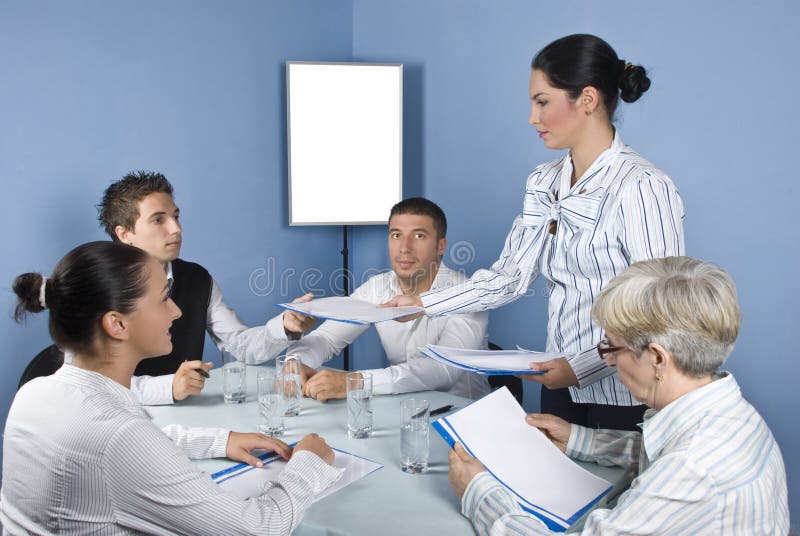 Group of business people at meeting table sitting on chair while a business young woman giving all folders with papers,blank chart for presentation in background,check also. Group of business people at meeting table sitting on chair while a business young woman giving all folders with papers,blank chart for presentation in background,check also