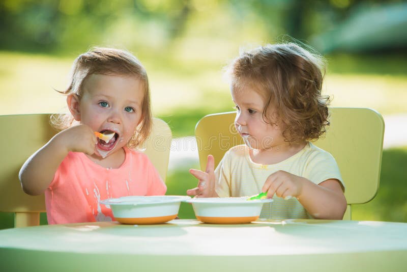 Two little 2 years old girls sitting at a table and eating together against a green lawn. Two little 2 years old girls sitting at a table and eating together against a green lawn