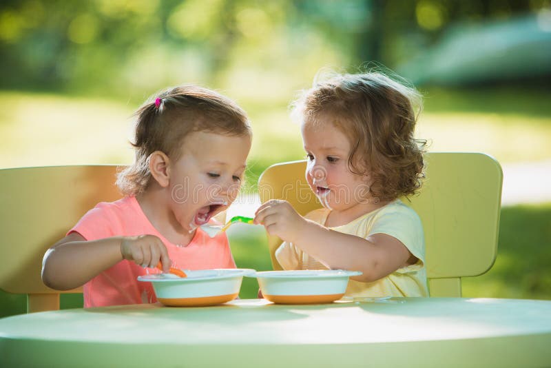 Two little 2 years old girls sitting at a table and eating together against a green lawn. Two little 2 years old girls sitting at a table and eating together against a green lawn