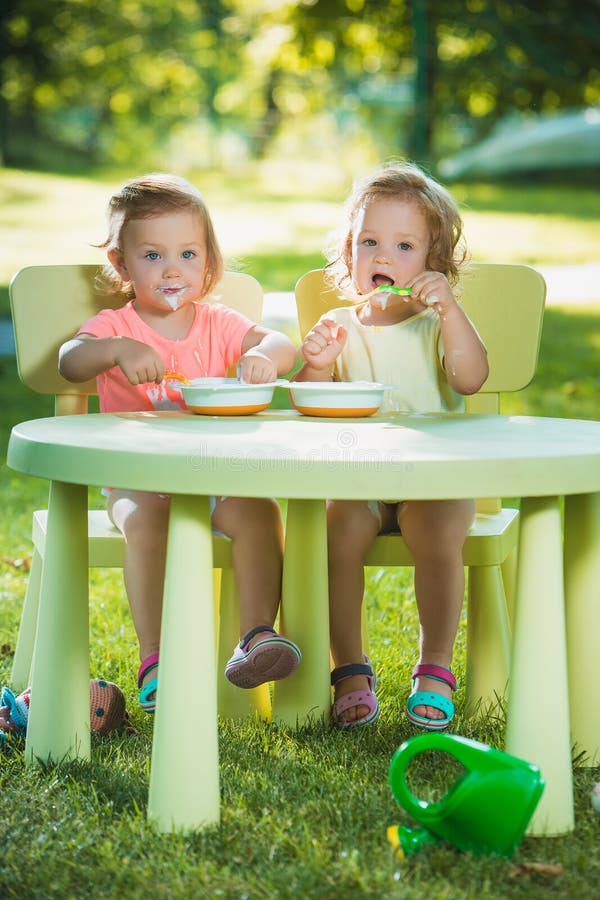 Two little 2 years old girls sitting at a table and eating together against a green lawn. Two little 2 years old girls sitting at a table and eating together against a green lawn