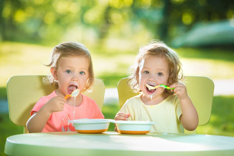 Two little 2 years old girls sitting at a table and eating together against a green lawn. Two little 2 years old girls sitting at a table and eating together against a green lawn