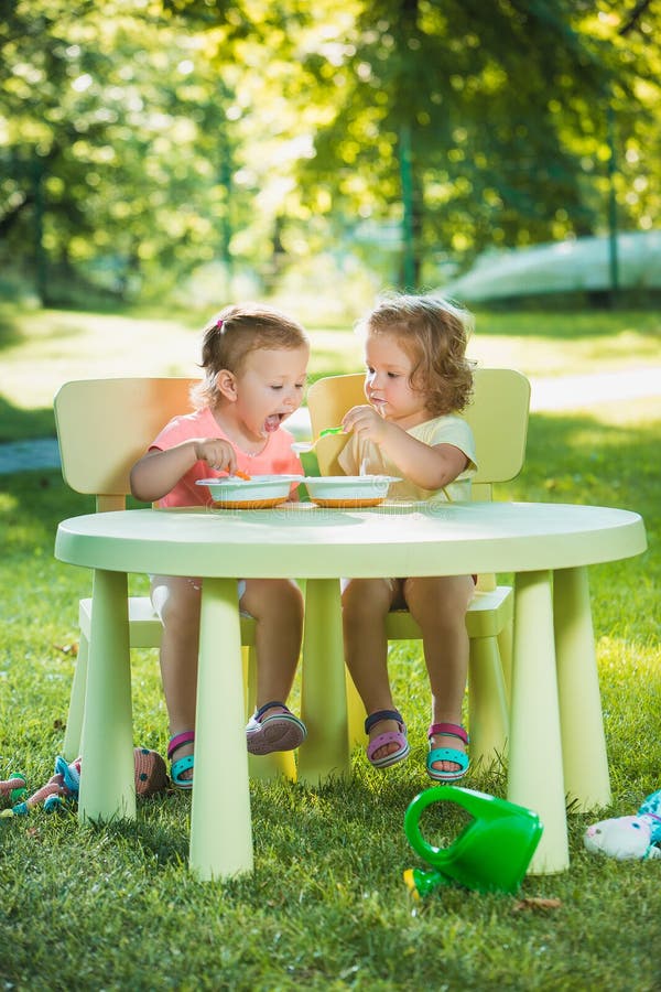 Two little 2 years old girls sitting at a table and eating together against a green lawn. Two little 2 years old girls sitting at a table and eating together against a green lawn