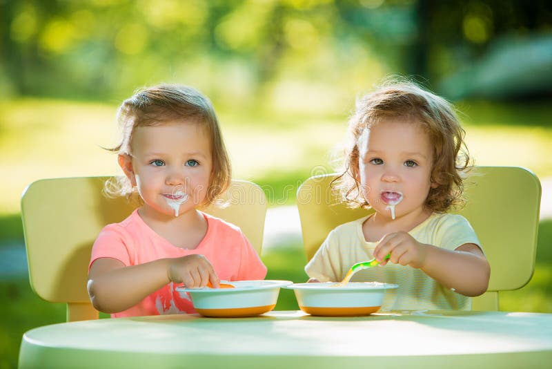 Two little 2 years old girls sitting at a table and eating together against a green lawn. Two little 2 years old girls sitting at a table and eating together against a green lawn