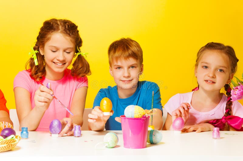 Boy and 2 girls painting Easter eggs at the table on the yellow background. Boy and 2 girls painting Easter eggs at the table on the yellow background