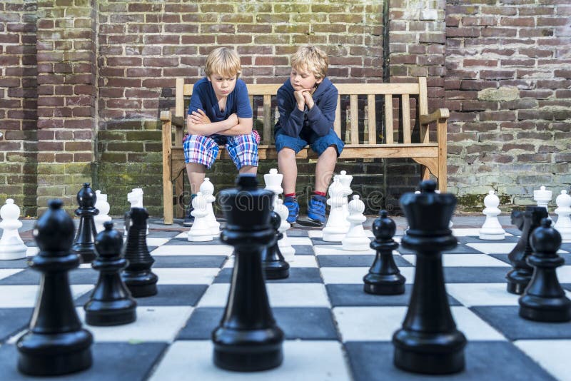 Two boys, sitting on a wooden bench, concentratedly thinking about their next move during an outdoors chess game with life sized pieces. Two boys, sitting on a wooden bench, concentratedly thinking about their next move during an outdoors chess game with life sized pieces.