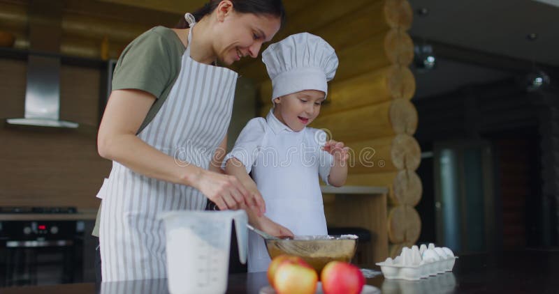 Мама в фартуке с сыном, одетым в шефов, одетых в одноразовый кашель для яблочного пирога на кухне. маленький шеф-повар акции видеоматериалы