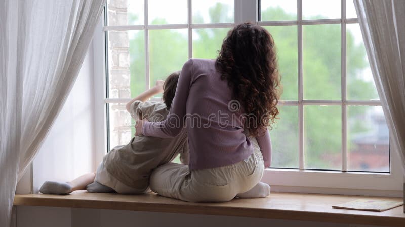 мама и сын сидят на подоконнике и смотрят в окно, обнимаясь друг с другом дома на карантин covid19