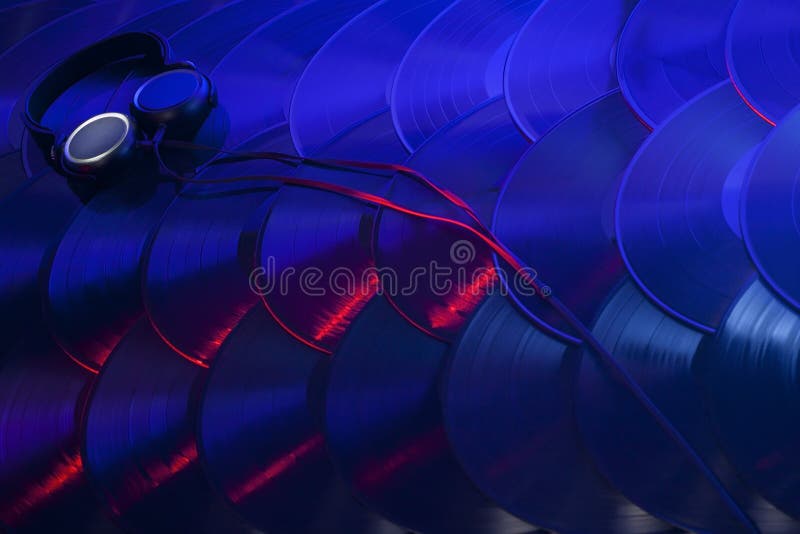 A pair of headphones sitting on several vinyl records overlapping in an organized array and lit with red, blue, purple and white lights. A pair of headphones sitting on several vinyl records overlapping in an organized array and lit with red, blue, purple and white lights.